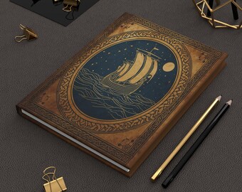 Peut inclure: Un journal à couverture rigide avec un design vintage. La couverture présente un ovale bleu avec une illustration dorée d'un navire viking naviguant sur la mer sous un ciel étoilé. Des crayons et des pinces à reliure dorés sont sur la surface gris foncé.