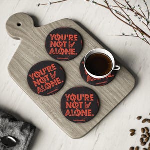Puede incluir: Cuatro posavasos negros con el texto "YOU'RE NOT ALONE" en rojo, colocados sobre una tabla de madera. Una taza blanca llena de líquido oscuro está sobre un posavasos. También se ven granos de café y una cuchara.
