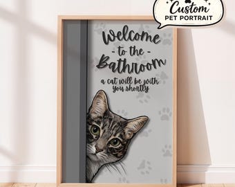 May include: A framed wall print featuring a gray tabby cat peeking from behind a door frame. The text reads: Welcome to the Bathroom, a cat will be with you shortly. The background is light gray with scattered dark gray paw print patterns.