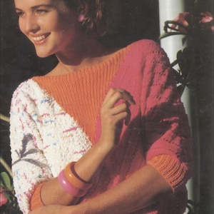 May include: A colour-blocked sweater in shades of orange, pink, and white. The sweater features a textured knit design with a wide neckline and long sleeves. A pink and orange bracelet is worn on the arm.