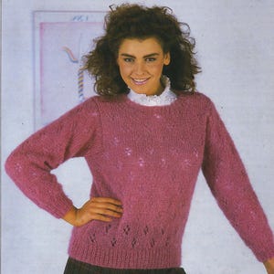 Może przedstawiać: Różowy dzianinowy sweter z okrągłym dekoltem i długimi rękawami. Sweter ma teksturowany wzór i jest noszony z białym kołnierzem z falbankami i spódnicą w kratę. Kobieta się uśmiecha.