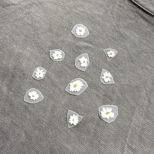 May include: A selection of daisy flower appliques with white petals and yellow centres. The appliques are on a grey fabric background, possibly a shirt or other garment. The appliques are of varying sizes and shapes.