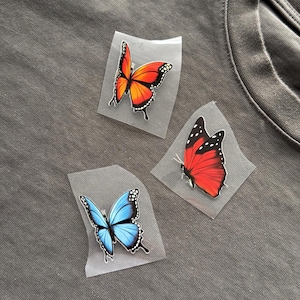 May include: Three butterfly iron-on patches in vibrant colours. One is orange and yellow, another is red, and the third is blue. Each butterfly has black detailing and is on a clear backing. The patches are on a grey fabric.