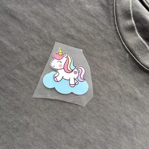 May include: A colorful unicorn iron-on patch. The unicorn has a pink mane and tail, a yellow horn, and is sitting on a blue cloud. The patch is on a gray t-shirt.