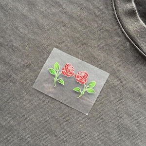May include: Two red rose iron-on patches with green leaves and stems are shown on a grey fabric background. The roses have detailed petals. The patches are on a clear backing.