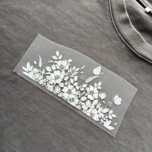 May include: A grey t-shirt featuring a floral and butterfly transfer. The design is white and grey, depicting detailed flowers, leaves, and butterflies. The shirt has a crew neck.