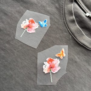 May include: Close-up of a grey t-shirt with two floral and butterfly patches. The patches feature watercolour-style flowers in pink and orange, and butterflies in blue and orange. The t-shirt has a crew neck.