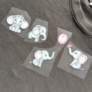 Op de afbeelding: Vier opstrijkbare stoffen patches met baby-olifanten in aquarel-stijl. De olifanten zijn lichtblauw met roze oren en accenten. Eén olifant heeft een bloem, een andere een roze ballon en een andere een vlinder.