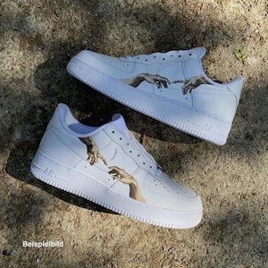 Pode incluir: Tênis brancos com um design personalizado com uma ilustração de mãos em estilo renascentista. Os sapatos têm um design clássico de cano baixo com solas e cadarços brancos. A imagem mostra os sapatos em uma superfície clara e texturizada.