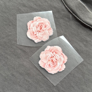 May include: Two pink flower iron-on patches on a grey fabric surface. Each patch features a detailed floral design with a light pink colour and a silver glitter outline. The patches are on clear, square backing.