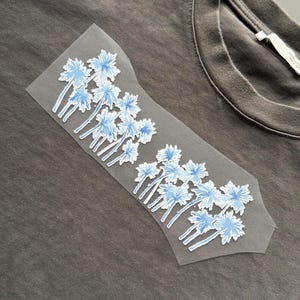 May include: A grey t-shirt with a floral design. The design features light blue flowers with white stems arranged in a decorative pattern. The shirt has a crew neck and a tag on the inside.