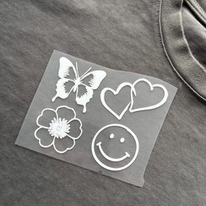 May include: A grey t-shirt with a white, transparent patch. The patch features a butterfly, two hearts, a flower, and a smiley face, all outlined in white. The design is simple and clean.