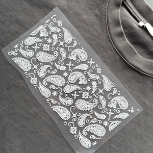 May include: A grey t-shirt with a white paisley design transfer sheet. The paisley pattern features intricate floral and teardrop shapes. The shirt has a crew neck and short sleeves.