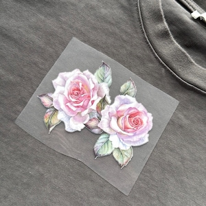 May include: A grey t-shirt with a floral design. The design is a watercolour illustration of two pink roses with green leaves, applied to a transparent square.