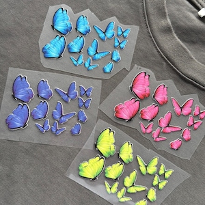May include: Close-up of a gray t-shirt with four sets of butterfly iron-on transfers. The butterflies are in shades of blue, pink, and yellow-green. The transfers are on clear backing.