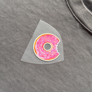 May include: A grey t-shirt featuring a pink donut graphic. The donut has pink icing, colourful sprinkles, and a bite taken out of it. The graphic is partially covered by a transparent material.