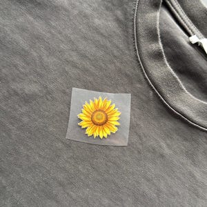 May include: A gray t-shirt with a yellow sunflower graphic on the chest. The sunflower has yellow petals and a brown center. The graphic is on a clear, square backing.