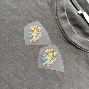 May include: Close-up of a grey t-shirt with two cherub patches. Each patch shows a cherub with golden wings, blue cloth, and a bouquet of flowers. The patches are on a semi-transparent background.