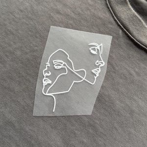 May include: A grey t-shirt featuring a white, continuous line drawing of two faces. The design shows the profiles of two people in a minimalist style. The t-shirt has a crew neck and is made of a soft, textured fabric.
