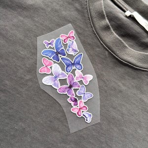 May include: A gray t-shirt with a cluster of colorful butterfly decals. The butterflies are in shades of pink, purple, and blue, with intricate wing details. The decals are arranged on a clear, irregular-shaped backing.