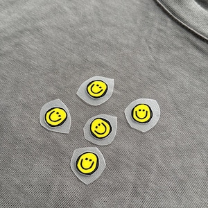 May include: Five yellow smiley face iron-on patches with black outlines and a clear backing are arranged on a grey fabric surface. The patches are in a diamond-like pattern. The smiley faces have a simple, cheerful design.