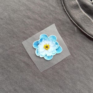 May include: A close-up of a gray t-shirt with a light blue and white flower patch. The flower has a yellow center and is attached to a square of sheer fabric. The t-shirt is made of a soft, textured material.