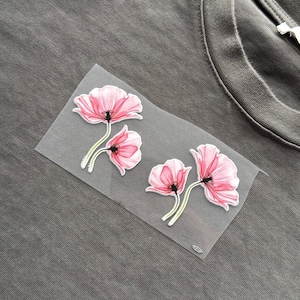 May include: A grey t-shirt with a floral design featuring three pink poppy flowers with green stems and black centres. The flowers are attached to a clear backing. The t-shirt fabric has a textured appearance.