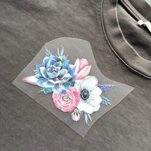 May include: A gray t-shirt featuring a floral design. The design includes a blue succulent, pink tulips, a white anemone, and other pink and purple flowers. The flowers are arranged on a transparent patch.