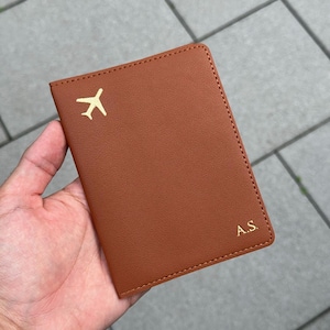 Può includere: Un porta passaporto in pelle marrone con un'icona di aeroplano dorata e le iniziali "A.S.". La copertina è liscia e ha bordi cuciti. Un accessorio da viaggio.