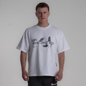 Könnte beinhalten: Weißes T-Shirt mit kurzen Ärmeln und dem grauen Schriftzug "UNRESTRICTED" sowie einer Grafik von fliegenden Vögeln. Das T-Shirt wird von einer Person getragen.