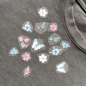 May include: A grey t-shirt decorated with various floral and butterfly appliques. The appliques showcase delicate watercolour designs in pink, blue, and white. The t-shirt's fabric has a slightly worn appearance.