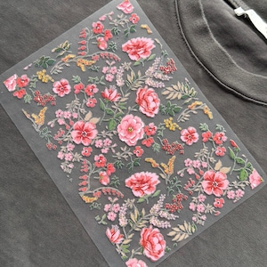 May include: A floral transfer sheet featuring pink and red flowers, green leaves, and yellow details. The sheet is positioned on a grey t-shirt, demonstrating its use. The flowers are intricately designed and arranged in a decorative pattern, ready for application.