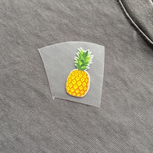 May include: A small, colourful pineapple iron-on patch with a yellow body and green leaves is attached to grey fabric. The patch is on a transparent backing. The fabric appears to be a t-shirt or similar garment.