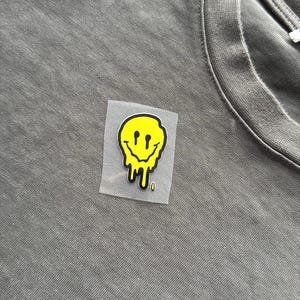 May include: Close-up of a grey t-shirt featuring a yellow melting smiley face graphic. The smiley face has black eyes and a dripping mouth. The graphic is on a clear, rectangular backing.