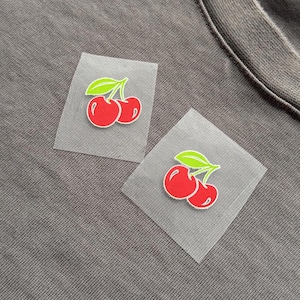 May include: Two iron-on cherry patches. Each patch features a pair of red cherries with white highlights and green leaves. The patches are on a transparent backing, set against a grey fabric background.