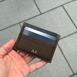 Puede incluir: Un tarjetero de cuero marrón con las iniciales "A.S." en letras doradas. El tarjetero está lleno de varias tarjetas. Las costuras son blancas. El cuero tiene un aspecto texturizado.