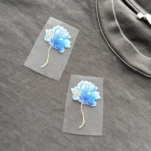 May include: Two blue floral iron-on patches on a grey t-shirt. The flowers have a watercolour effect with shades of blue and white. The patches are rectangular with a transparent background and a yellow-green stem.