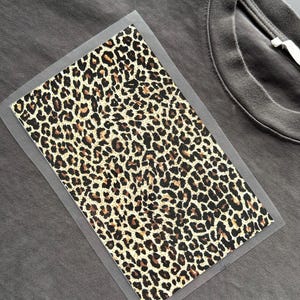 Puede incluir: Una camiseta gris oscuro con un parche con estampado de leopardo. El parche es rectangular con un borde transparente y presenta un patrón de manchas marrones y negras sobre un fondo beige. La camiseta tiene cuello redondo.