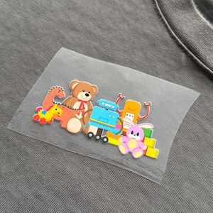May include: A gray t-shirt with a colorful patch featuring cartoon toys. The patch includes a brown teddy bear, a red and orange dinosaur, a blue robot, a yellow robot, and a pink bunny. The toys are arranged on a clear, rectangular patch.