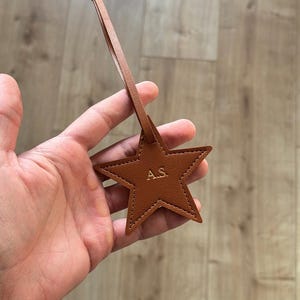 May include: A brown leather star-shaped tag with the initials "A.S." embossed in gold. The tag is attached to a thin leather strap with a knotted end. The tag is held in a person's hand.