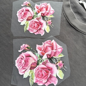 May include: Two floral iron-on patches with watercolour-style pink roses, green leaves and buds. The patches are on a grey background, possibly a shirt or fabric.
