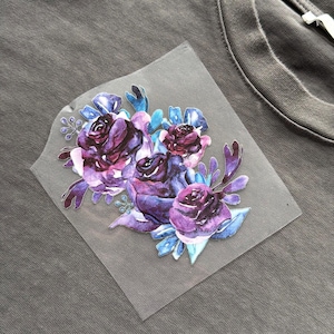 May include: A grey t-shirt with a floral design. The design features watercolour roses in shades of purple and blue, with green leaves. The floral graphic is on a clear, square patch.