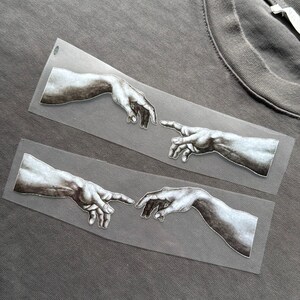 May include: Grey t-shirt with two rectangular patches depicting a black and white illustration of hands reaching out, inspired by Michelangelo's "The Creation of Adam". The shirt has a crew neck.