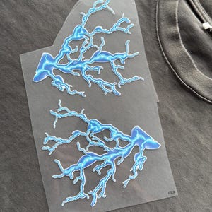 Könnte beinhalten: Zwei blau-weiße Blitz-Aufnäher auf einem grauen T-Shirt. Die Aufnäher befinden sich auf einer transparenten Trägerfolie und sind zum Aufbügeln bereit. Die Blitze haben ein detailliertes, verzweigtes Design.