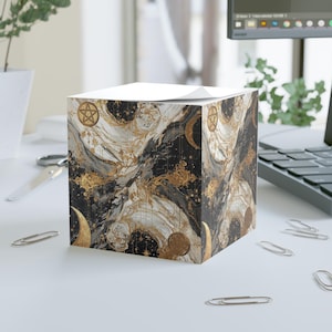 Peut inclure: Un distributeur de blocs-notes en forme de cube avec un motif céleste. Le motif présente des lunes, des étoiles et des pentagrammes dorés sur un fond marbré noir et blanc. Le dessus du cube contient une pile de papier blanc.