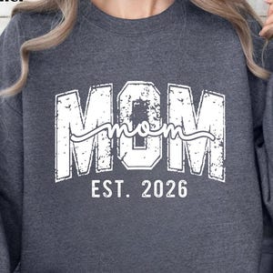 May include: Dark gray heather sweatshirt with the word "MOM" in large, distressed white letters, with "mom" in cursive script above "EST. 2026". The sweatshirt is a crew neck style.