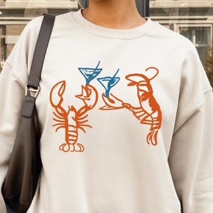 May include: A cream-colored sweatshirt featuring an orange lobster design holding two blue cocktails. The design is centered on the front of the sweatshirt. The person is wearing a pleated skirt and holding a white coffee cup.