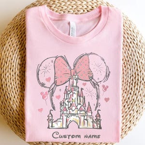 A pink t-shirt with a drawing of a castle and two pink bows, along with the text "Custom Name" printed on it.