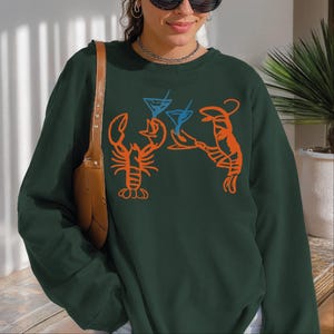 May include: Dark green sweatshirt featuring orange lobster and blue cocktail glass graphics. The design includes two lobsters holding up cocktail glasses. A fun, casual top for seafood lovers.