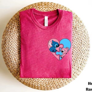 A pink t-shirt with a heart-shaped graphic featuring two cartoon characters, one blue and one pink, is displayed on a woven basket.